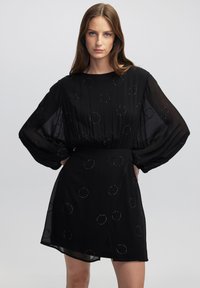 Schwarzes Kleid aus durchscheinendem Stoff mit kreisförmigen Mustern und Perlenbesatz. Lange, luftige Ärmel und eine lockere Passform, mit einem leichten Schlitz.