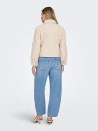Kvinde med langt hår iført en beige fleecejakke, lyseblå viddebenede jeans og beige slingback-sko med lav hæl, set bagfra.