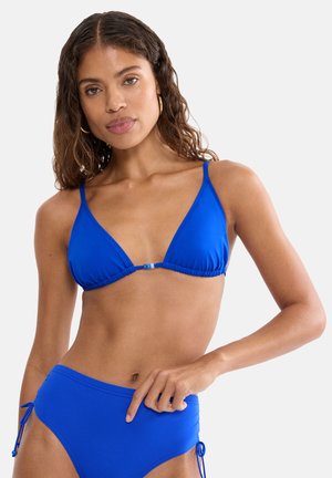 Frau mit langen, welligen Haaren, die einen knallblauen Bikini mit verstellbaren seitlichen Bändern trägt und mit einer Hand auf der Hüfte posiert.