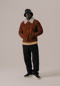 Chaqueta de ante marrón con un cuello de borrego gris, camiseta de cuello redondo beige, pantalones de chándal negros y zapatillas negras. El modelo lleva un sombrero de cubo negro.