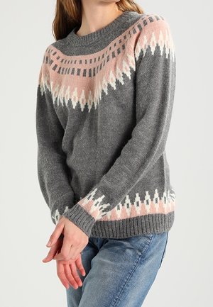 Femme portant un pull en tricot gris avec des motifs géométriques roses et blancs sur la poitrine, les manches et l'ourlet, associé à un jean bleu.