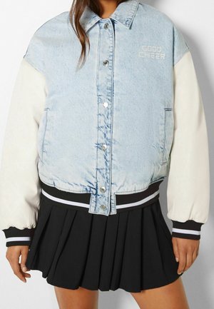 Persoon draagt een lichtblauwe spijkerjas met witte mouwen, tekst "GOOD CHEER" op de borst, en een zwarte geplooide rok.