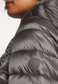 Graue Daunenjacke aus glänzendem Stoff, mit gestepptem Muster, rundem schwarzem Logo-Patch am Oberarm und hochgeschlossenem Design.