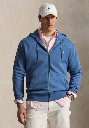 Mężczyzna w białej czapce, niebieskiej bluzie na zamek z kapturem na różowej koszuli w paski, białych spodniach i zegarku, stojący z jedną ręką w kieszeni.