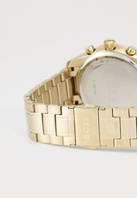 Orologio in acciaio inossidabile dorato con bracciale a maglie, presenta una cassa tonda, tre pulsanti e il logo "BOSS" incisisul fermaglio.