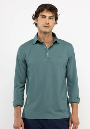 Hombre con cabello rizado oscuro que lleva una camiseta polo de manga larga color verde azulado con dos botones y mangas remangadas sobre un fondo blanco liso.
