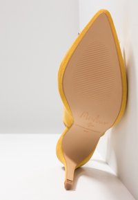 Chaussure à talon haut en daim jaune, positionnée avec la semelle vers le haut contre une surface blanche, montrant un bout pointu et un talon fin.