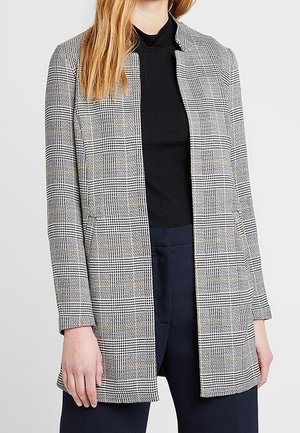 Vrouw draagt een grijze geruite blazer over een zwarte coltrui en donkerblauwe broek tegen een effen lichte achtergrond.