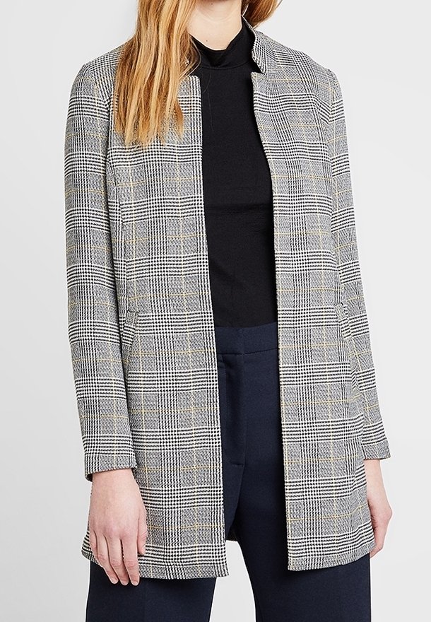 Femme portant un blazer à carreaux gris sur un col roulé noir et un pantalon bleu marine foncé sur un fond clair uni.