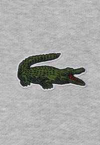 Écusson crocodile vert brodé avec bouche ouverte et langue rouge sur fond de tissu gris clair.
