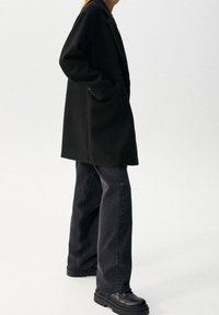 Cappotto oversized nero con un design sartoriale, dotato di bottoni davanti e tasche laterali. Abbinato a jeans scuri a vita alta e scarpe nere massicce.