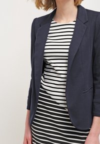 Marinblå kavaj med en strukturerad design och spetsiga slag, bärs över en svart-och-vit randig skjorta. Tyget verkar slätt och åtsittande.