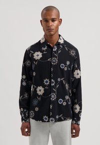 Camisa negra con estampado floral y flores bordadas en blanco, azul y gris. Cuenta con cuello abotonado y mangas largas. Tejido texturizado.