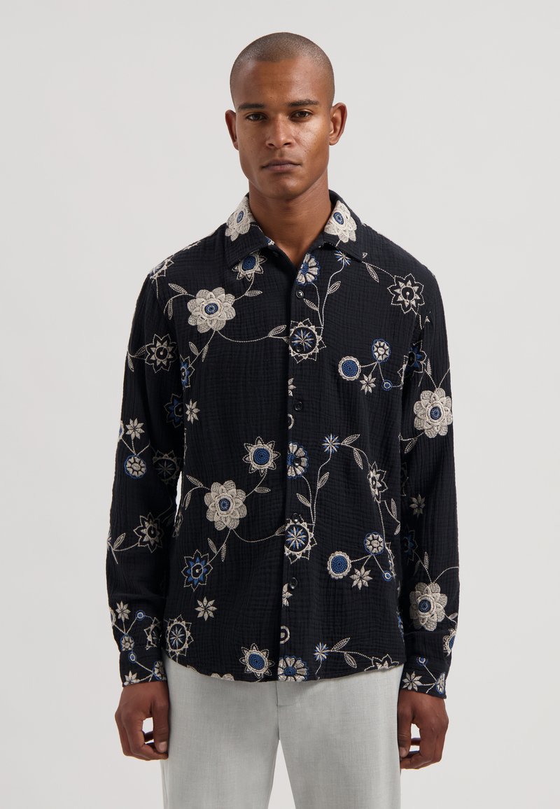 Camisa negra con estampado floral y flores bordadas en blanco, azul y gris. Cuenta con cuello abotonado y mangas largas. Tejido texturizado.