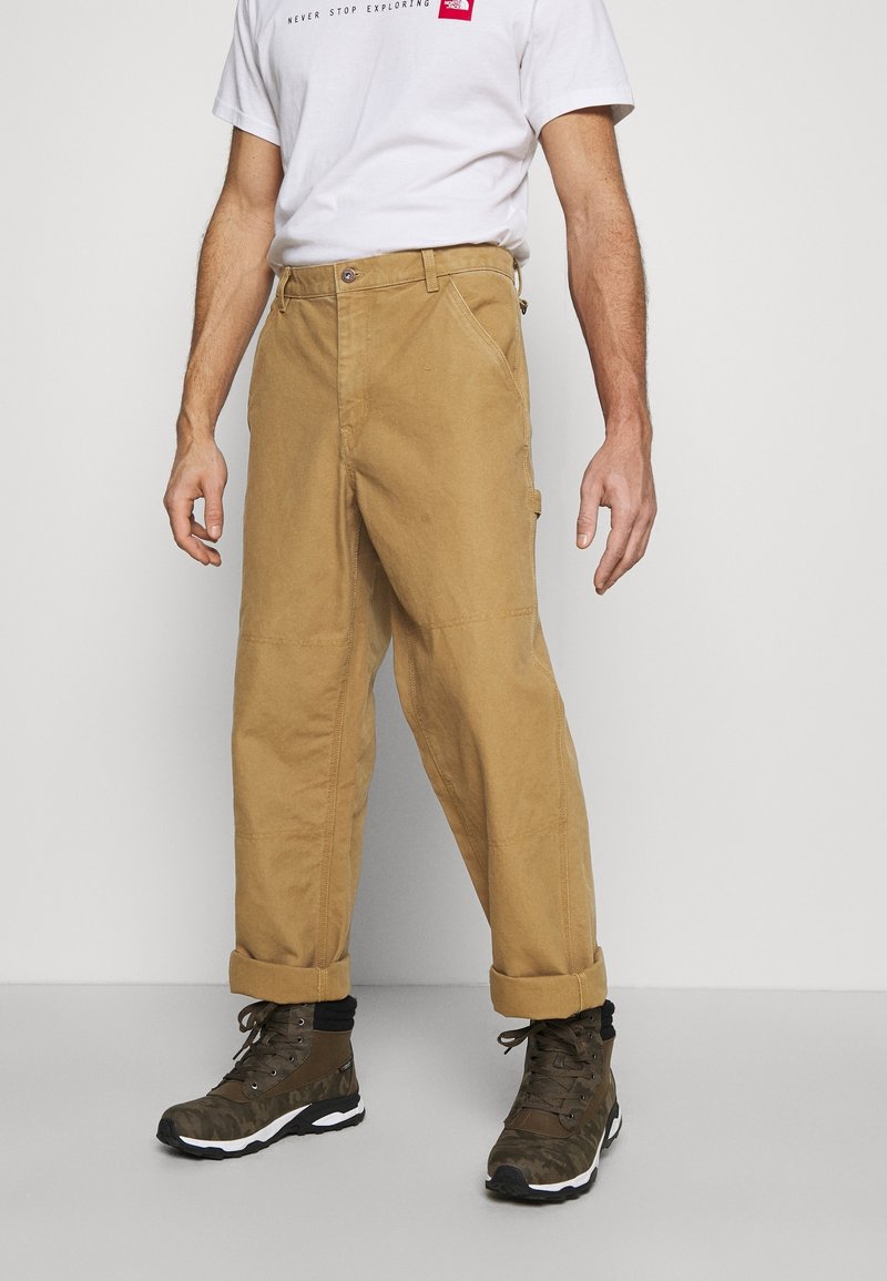 Homme portant un pantalon de travail beige retroussé, un t-shirt blanc avec un logo rouge et des bottes de randonnée vert foncé, debout devant un fond uni.