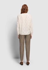 Vrouw met lang rood haar, gekleed in een crèmekleurige blouse met oogjes, bruine geruite broek en schoenen met luipaardprint, staand met de rug naar de camera op een grijze achtergrond.