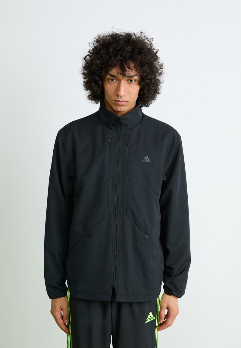 Ung person med krøllet hår iført en sort Adidas-jakke og matchende sorte Adidas-bukser med neongrønne striber, stående foran en ensfarvet hvid baggrund.