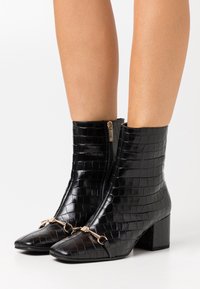 Botas de tobillo de piel sintética de cocodrilo en negro, con punta afilada, detalles en dorado y cremallera lateral, sobre un tacón grueso.