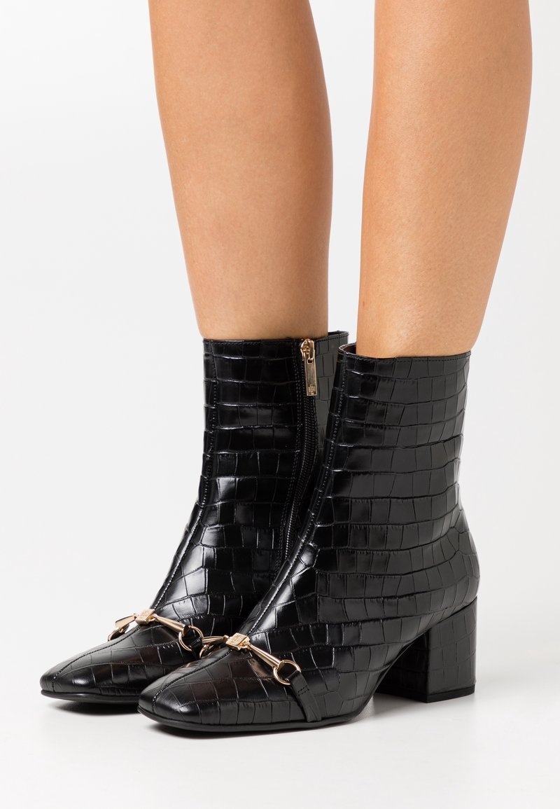 Botas de tobillo de piel sintética de cocodrilo en negro, con punta afilada, detalles en dorado y cremallera lateral, sobre un tacón grueso.