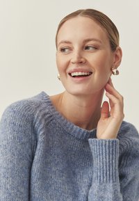 Pull en tricot bleu clair avec un col rond et des poignets côtelés, présentant une finition douce et texturée. Le modèle porte des boucles d'oreilles en or.
