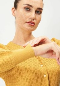 Senapsgul stickad cardigan med strukturerat mönster, ribbade ärmslut och rund halsringning, med tre knappar framtill.