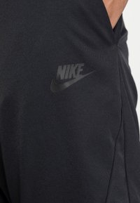 Black athletic pants made of textured fabric, featuring a subtle Nike logo in a glossy finish on the left thigh.
