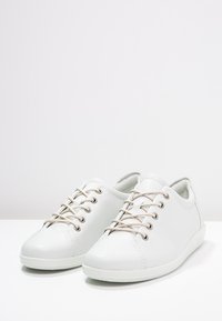 Baskets basses en cuir blanc avec lacets beige, œillets métalliques et semelles en caoutchouc, présentées sur une surface blanche sur fond clair.