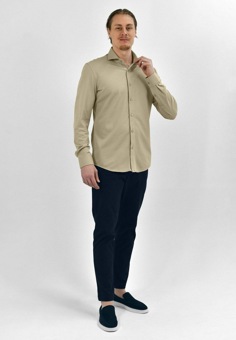 Beige langwerpige overhemdd met knopen en een kraag, gecombineerd met donkere broeken en zwarte instapschoenen, weergegeven op een staand model.