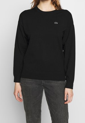 Frau trägt ein schwarzes Sweatshirt mit langen Ärmeln und kleinem weißem Krokodil-Logo auf der linken Brust sowie dunkelgraue Jeans.