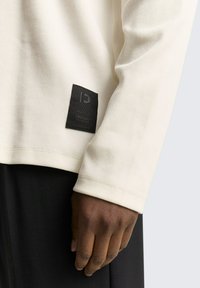 Crème lange mouwen shirt met een geribbelde textuur, voorzien van een zwart geweven label op de mouw en een ontspannen pasvorm. Gecombineerd met zwarte broek.