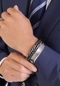 Metalen armband met onderling verbonden zwarte ketting en een glad zilveren paneel, gedragen om een pols, in contrast met een donkerblauwe jas en gestreepte stropdas.
