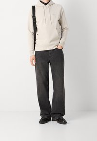 Felpa leggera color beige con tasca frontale, abbinata a jeans larghi grigi scuri e scarpe nere. Design semplice e minimalista.