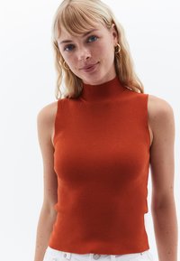 Top smanicato in maglia color ruggine con colletto alto e texture a coste, caratterizzato da una silhouette aderente e spaccature laterali per una maggiore libertà di movimento.