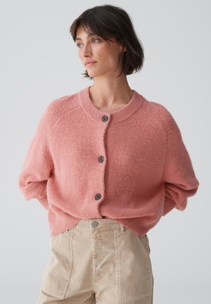 Soft pink knitted cardigan with a round collar, featuring five buttons and loose sleeves, paired with beige high-waisted trousers.