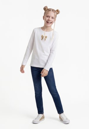 Lächelndes Mädchen mit geflochtenem Haar in zwei Dutts, trägt ein weißes Langarmshirt mit goldener Schleife, dunkle Jeans und weiße Sneaker, steht vor weißem Hintergrund.