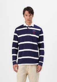 T-shirt de rugby à manches longues bleu marine avec des rayures horizontales blanches, un col, et des détails graphiques rouges. Matière en coton, coupe décontractée.