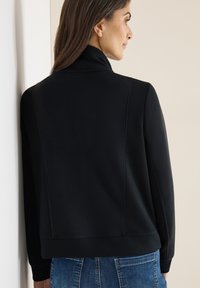 Černá sportovní bunda s vysokým límcem, žebrovanými detaily na ramenou a hladkou texturou. Kombinována se světle modrými džínami.