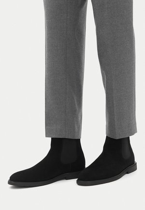 Schwarze Chelsea-Stiefel aus Wildleder, getragen zu grauen, maßgeschneiderten Hosen, die klare Linien und einen minimalistischen Stil zeigen.