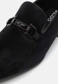Chaussure slip-on en velours côtelé noir avec un bout rond. Présente un accent en métal noir brillant avec des formes géométriques. Doublure intérieure lisse.