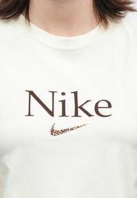 Biela košeľa s veľkým hnedým nápisom "Nike" a logom Nike swoosh so levanduľovým vzorom, umiestneným na strede hrudníka.