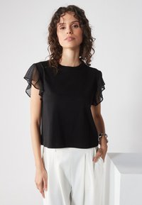 Haut noir à manches courtes avec des accents en tulle froncés, encolure ronde et coupe décontractée. Stylistiquement associé à un pantalon blanc plissé.