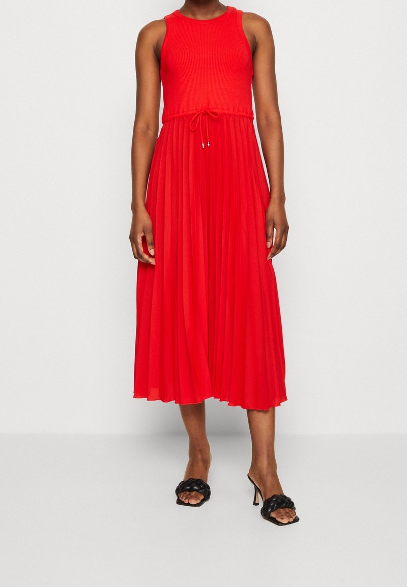 Rode, geribbelde tankjurk met een geplooide rok, een taille met trekkoord. Draagt zwarte sandalen met hak.