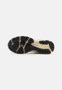 Semelle d'une chaussure de course beige et noire avec un motif de bande de roulement texturé pour adhérence, présentée sur un fond blanc uni.