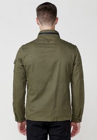 Olīvu zaļa, viegla veste ar stāvošu apkakli, pogu aprocēm un sānu kabatām. Satur redzamās šuves un logotipa detaļu aizmugurē.