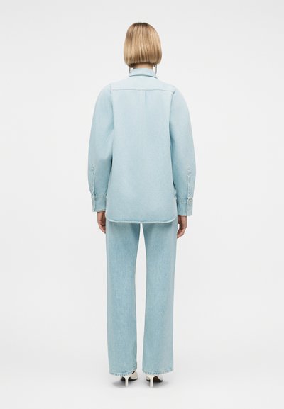 Γυναίκα με κοντά ξανθά μαλλιά που φοράει ανοιχτό μπλε πουκάμισο από τζιν και τζιν παντελόνι με φαρδιά μπατζάκια, στέκεται με την πλάτη στραμμένη προς τα εμπρός σε λευκό φόντο.