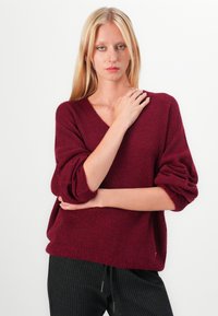Bordeaux v-hals sweater med løse, lange ærmer. Blød tekstur, afslappet pasform og en subtil guldaccent ved sømmen. Sæt sammen med stribede bukser.