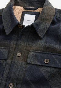 Kockovaný flanelový košeľa v tmavomodrých a hnedých tónach, s jemným béžovým fleece golierom, dvoma vreckami na hrudi a hnedými gombíkmi.