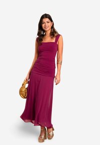 Lila gerafftes Kleid mit quadratischem Ausschnitt, schwingendem Rock und breiten Trägern. Kombiniert mit goldenen Plateauschuhen und einer goldenen Handtasche.