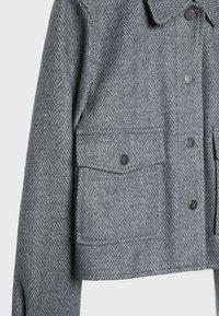 Szary żakiet w wzór rybiej ości o skróconym kroju, z dwiema przednimi kieszeniami i czarnymi zapięciami na napy. Wykonany z teksturowanej tkaniny o klasycznym wzorze.