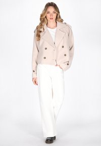 Afskåret beige dobbeltradet jakke med store knapper, brede slag og skulderepauleter, kombineret med hvide vide bukser og sorte sko.
